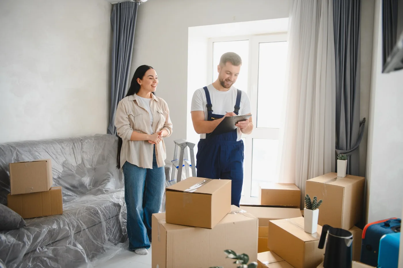 A mover writing on a clipboard during an in-home survey while a customer looks on, beside a wrapped sofa and packed boxes